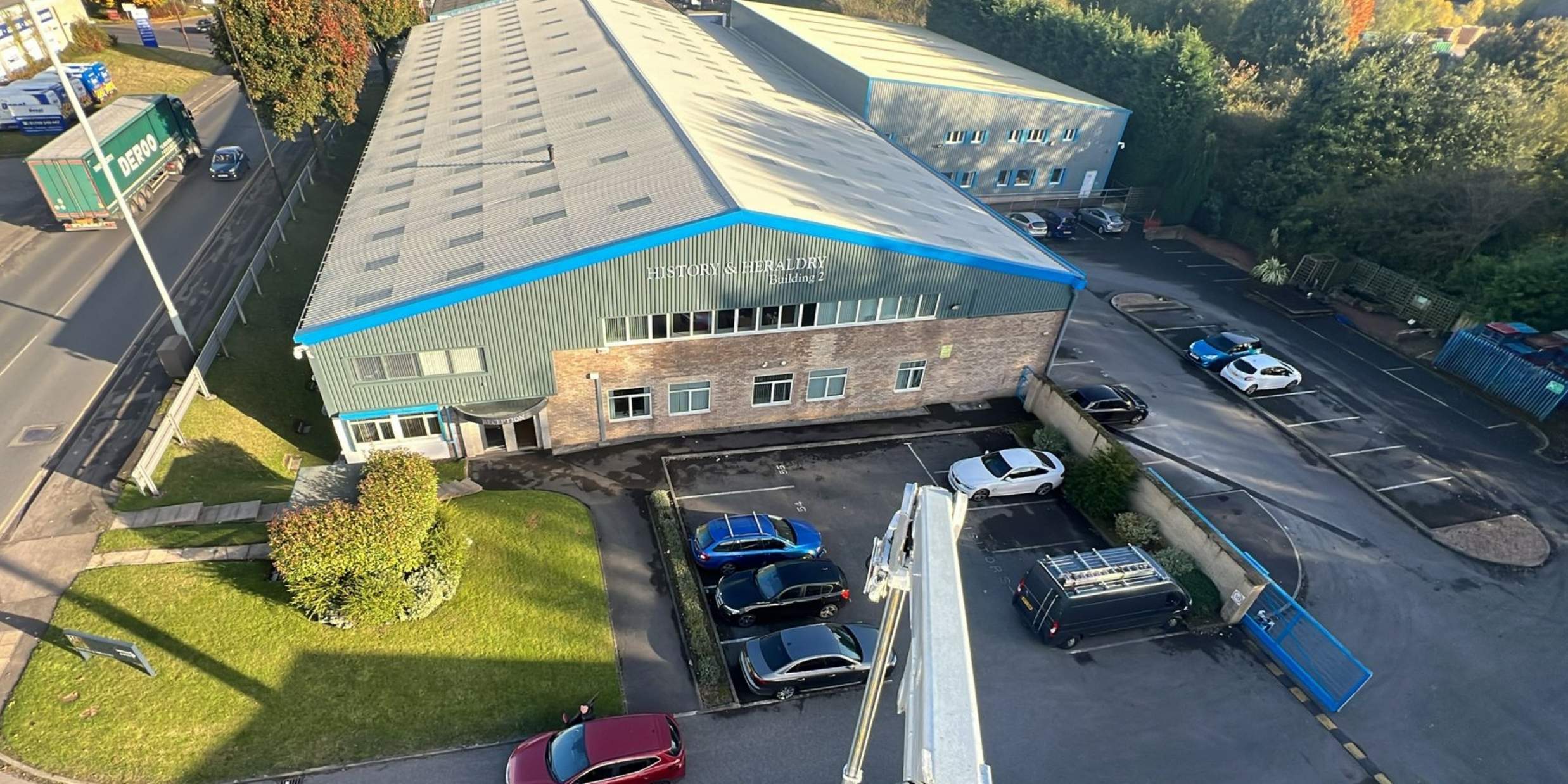 Aerial view of the History and Heraldry industrial unit in Sheffield, showing its profiled metal roof and resprayed blue-grey cladding, with a mobile elevating work platform parked alongside